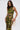 A woman with long brown hair wears the SEXY CAN I MOCKNECK BLOUSE WITH CUTOUT in green and yellow abstract patterns, paired with a matching high-slit skirt, and holds a gold fringed clutch against a plain white background.