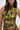 A woman wears the SEXY CAN I MOCKNECK BLOUSE WITH CUTOUT, a cropped sleeveless top in metallic green and gold with crisscross cutout waist details. She holds a small gold purse and has shoulder-length brown wavy hair.