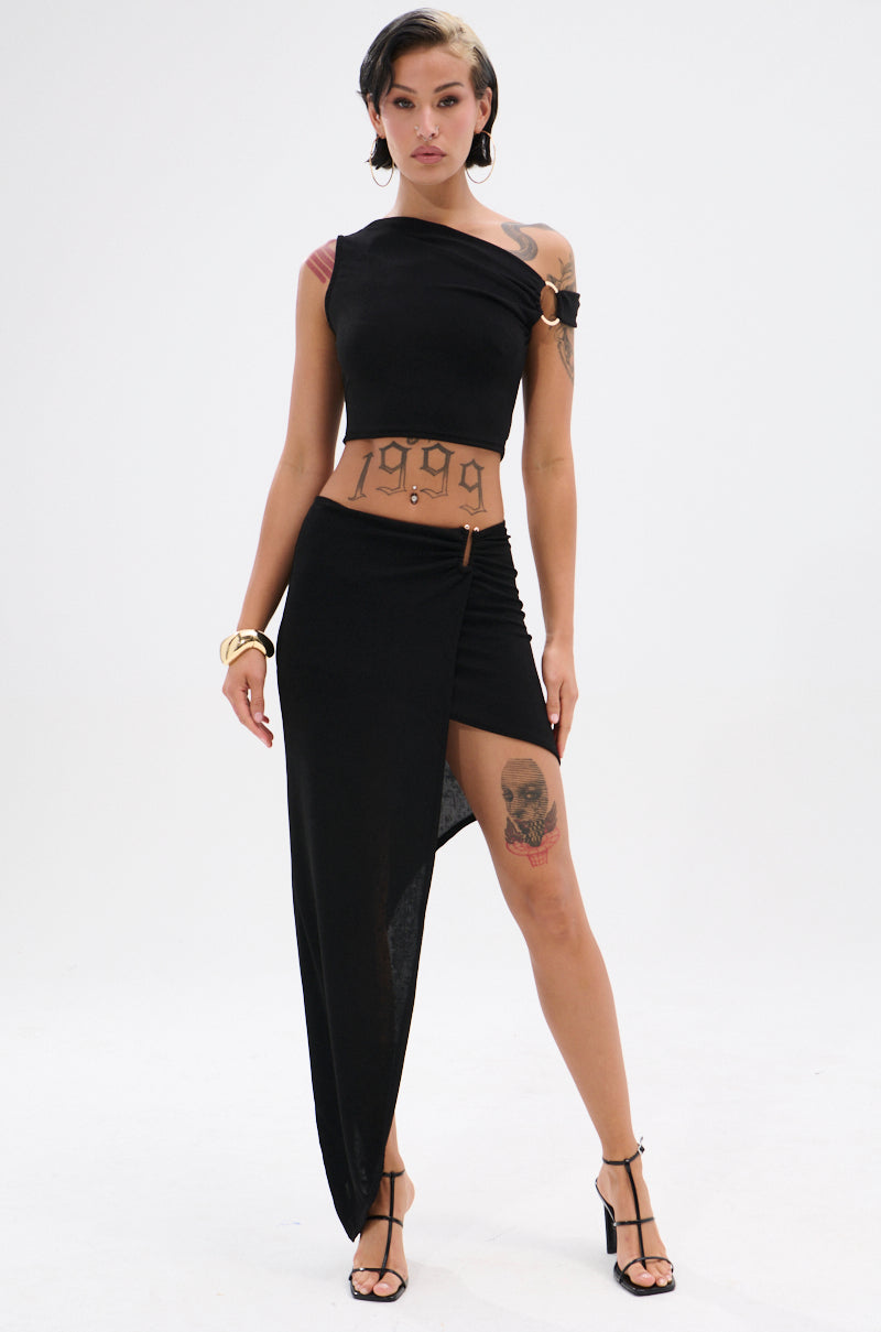 A person wearing a black asymmetrical crop top and the KYLIE SLINKY MIDI SKIRT with a ruched waistband and high-low hem stands confidently in black heels and gold jewelry, tattoos visible on torso and leg, against a plain white background.