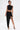 A person wearing a black asymmetrical crop top and the KYLIE SLINKY MIDI SKIRT with a ruched waistband and high-low hem stands confidently in black heels and gold jewelry, tattoos visible on torso and leg, against a plain white background.