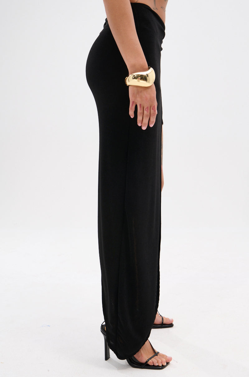 A person stands in profile wearing the KYLIE SLINKY MIDI SKIRT, styled with black high-heeled sandals and a large gold cuff bracelet against a plain white background.