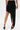 A person shown from the back against a plain white background wears the KYLIE SLINKY MIDI SKIRT, a black asymmetrical piece with a high-low hem—short on one side, long on the other—and black high-heeled sandals.