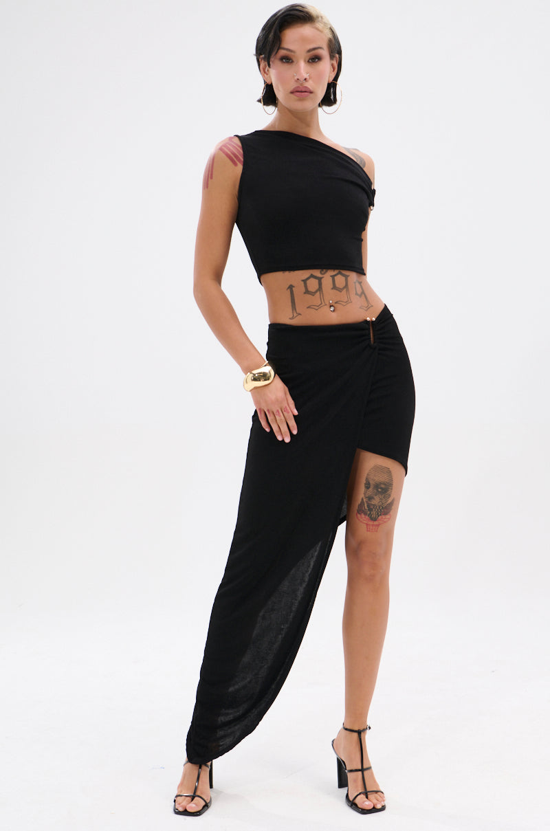 A model shows off visible tattoos as she pairs the KYLIE SLINKY MIDI SKIRT—featuring a ruched waistband and high-low hem—with a black one-shoulder crop top, strappy heels, and a gold bracelet for a chic asymmetrical look.