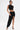 A model shows off visible tattoos as she pairs the KYLIE SLINKY MIDI SKIRT—featuring a ruched waistband and high-low hem—with a black one-shoulder crop top, strappy heels, and a gold bracelet for a chic asymmetrical look.