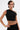 A person with short dark hair models the black KYLIE SLINKY OFF THE SHOULDER BLOUSE, styled with large hoop earrings and a gold bracelet, showing tattoos including "1999" on their stomach, against a plain background.