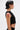 A person with short dark hair wears the black KYLIE SLINKY OFF THE SHOULDER BLOUSE and hoop earrings, revealing a red paint-drip shoulder tattoo and other tattoos on their forearm and side, standing in profile against a plain background.