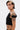 A person with short dark hair, hoop earrings, and tattoos wears the KYLIE SLINKY OFF THE SHOULDER BLOUSE in black stretch-knit, posing confidently against a plain white background.