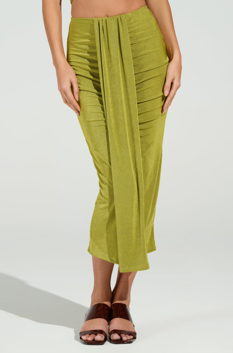 In an olive green VANJA SLINKY MIDI SKIRT with a curve-hugging silhouette and front drape, the person stands against a plain background. Brown slip-on sandals enhance the beach-ready look as their hands rest gracefully on the skirt.
