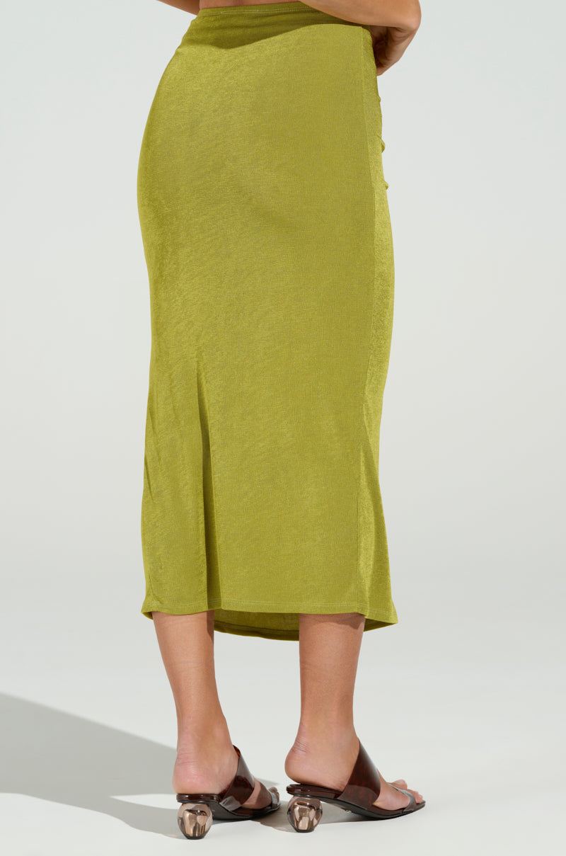 A person in a lime green Vanja Slinky Midi Skirt faces away, highlighting its body-contouring design, paired with brown slide sandals featuring transparent, distinct heels set against an off-white backdrop.