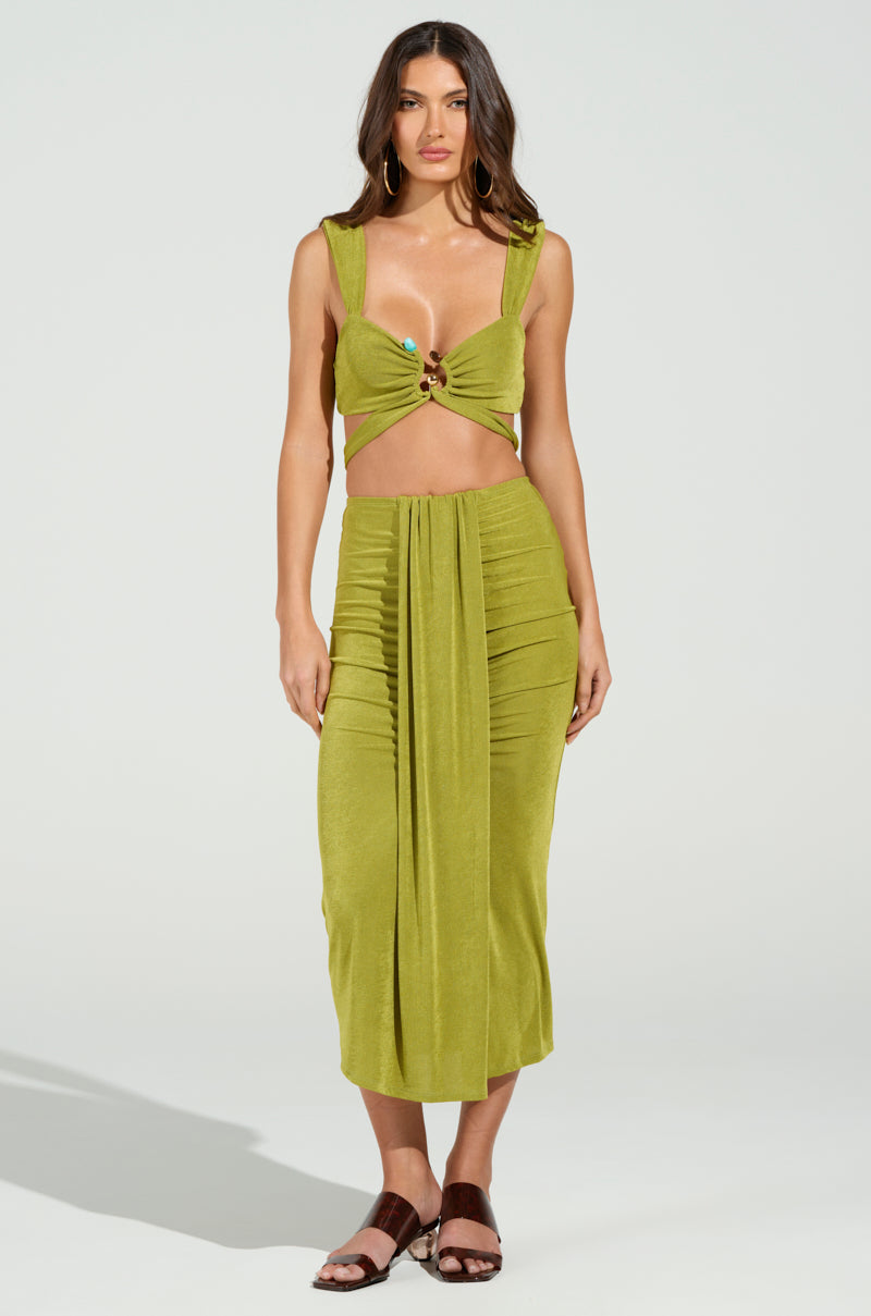 A person is modeling a beach ensemble with a green sleeveless cropped top featuring a knot detail and the VANJA SLINKY MIDI SKIRT with ruching, creating a curve-hugging silhouette that complements their brown sandals against a plain background.