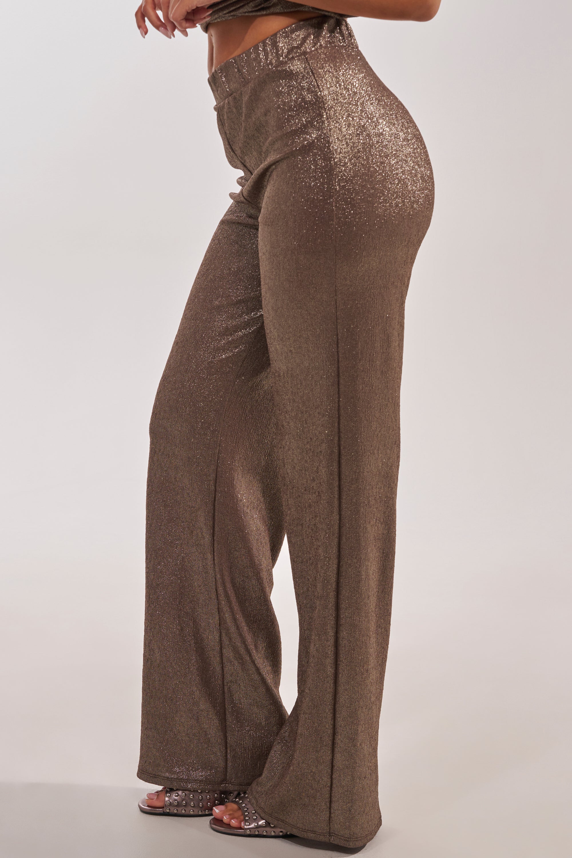 Wearing the HIGH STAKES RELAXED PANT, featuring a high-rise fit and shimmery wide-leg style, paired with matching open-toe heels. Only the lower half is shown against a plain light background. 1