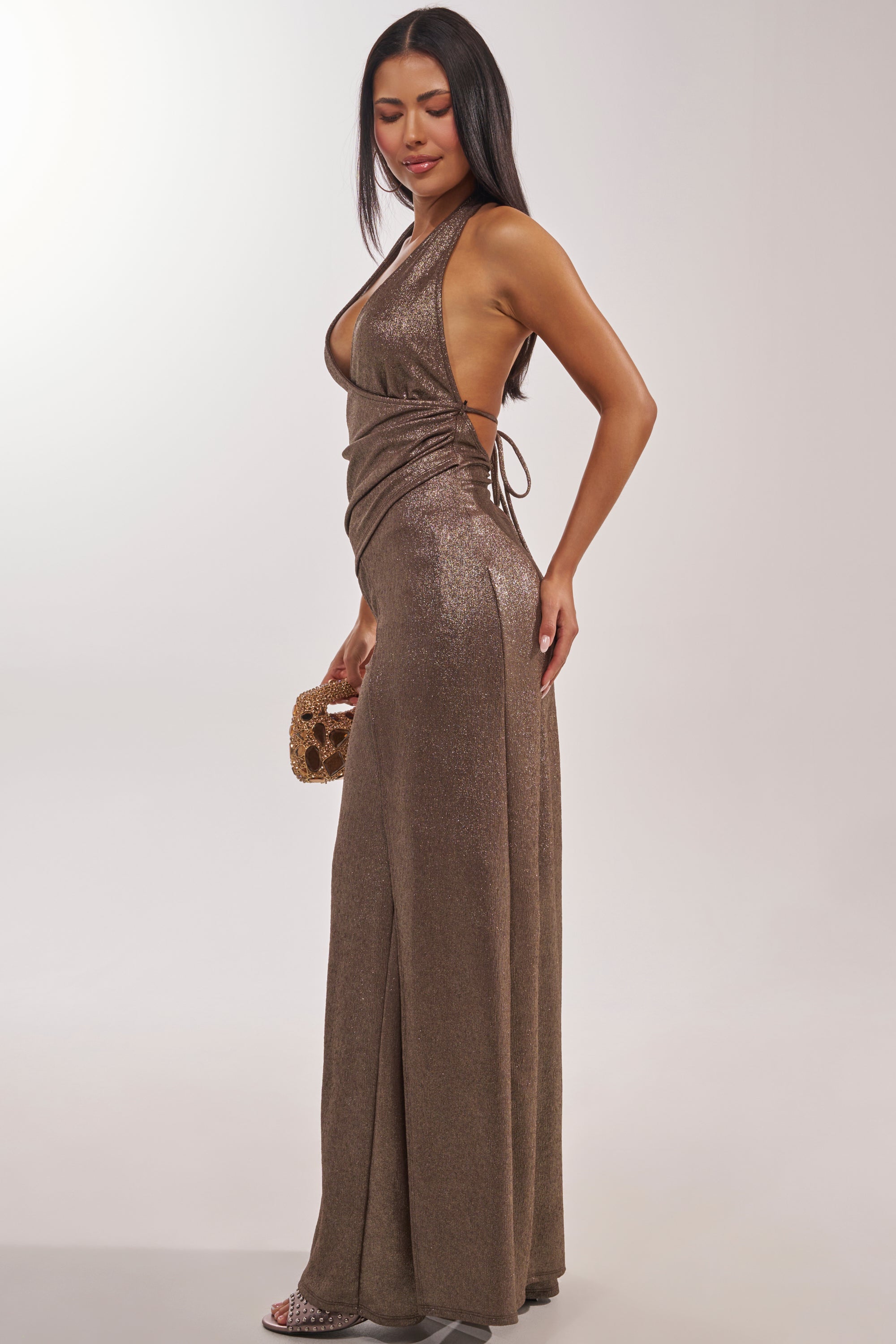 A woman poses in the HIGH STAKES HALTER BLOUSE, a metallic gold, backless top with a draped, sculpted silhouette and deep neckline. She pairs it with a small gold clutch and heeled sandals against a plain light background. 3