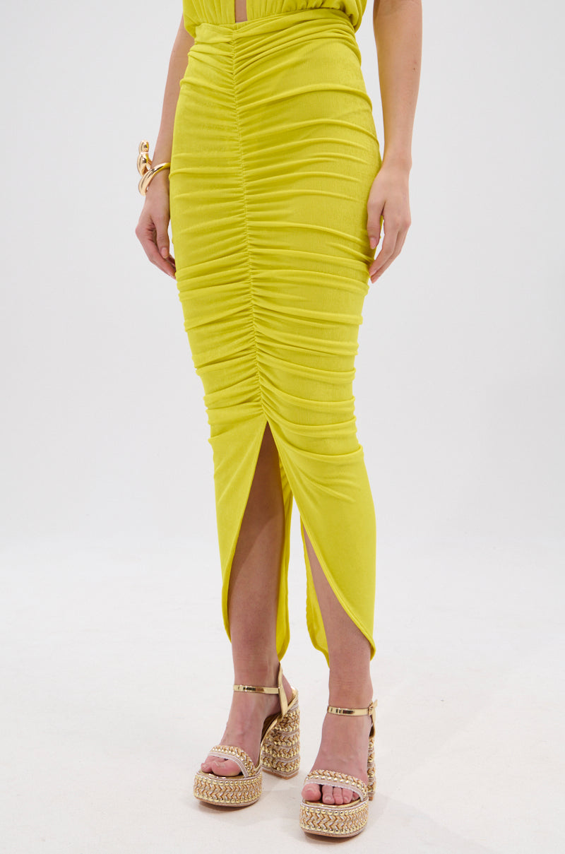 A person wearing the TIMELESS MESH MAXI SKIRT in bright yellow with ruching and a front slit, paired with platform sandals featuring woven details, stands against a plain white background. 2
