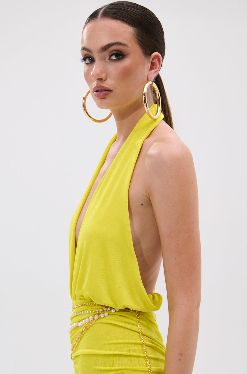 Woman in a sleeveless, deep v-neck yellow dress with gold hoops and a pearl chain belt, styled over the TIMELESS MESH HALTER NECK BODYSUIT, stands sideways against a white background with her dark hair in a sleek ponytail. 2