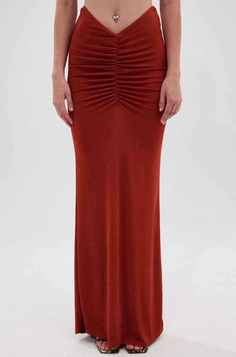 A person showcases the ON MY MIND SLINKY MAXI SKIRT in rust, featuring a ruched front and high-rise waist. They stand barefoot against a white background, with only the lower torso and legs visible. 1