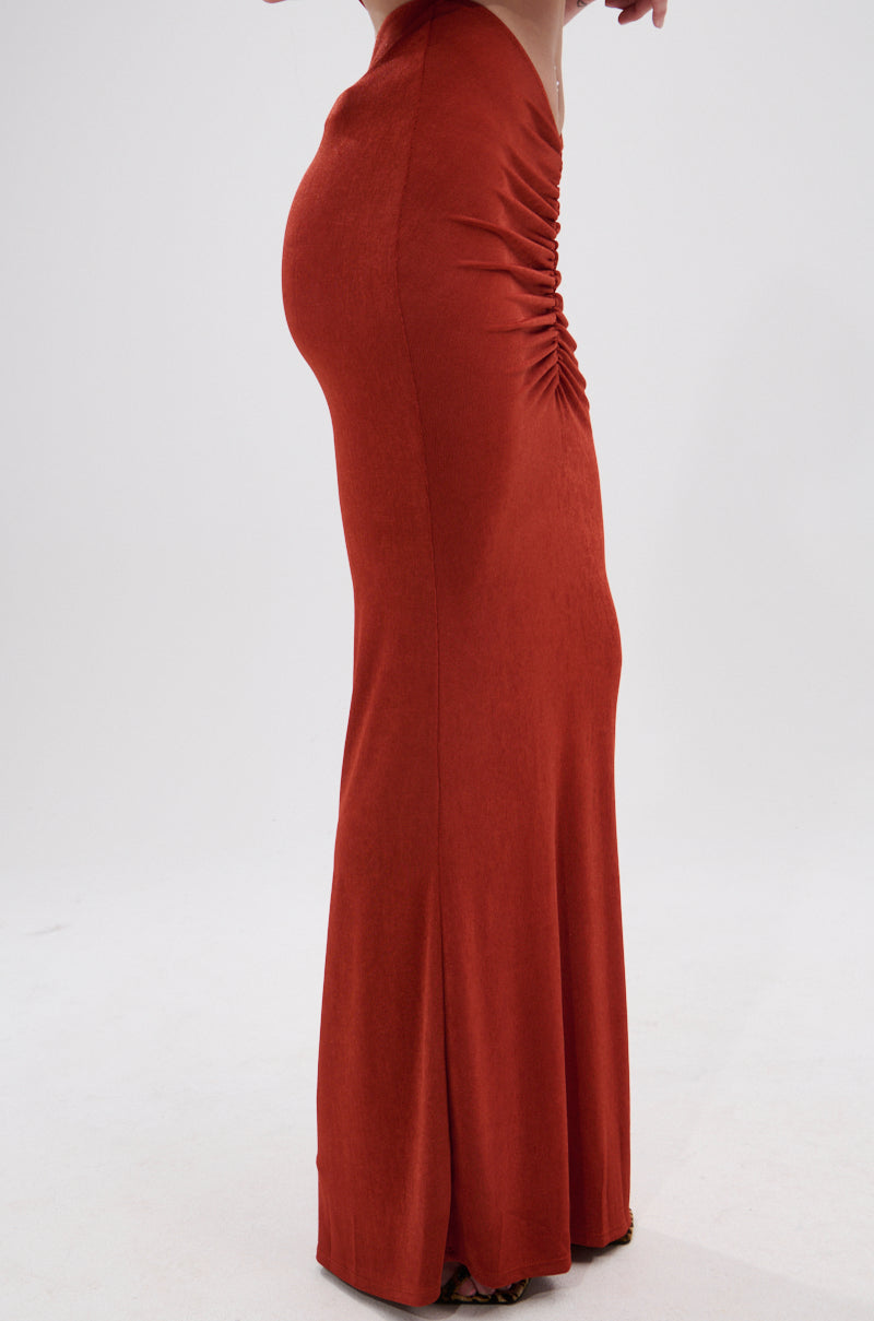 A person models the ON MY MIND SLINKY MAXI SKIRT in rust, featuring a fitted silhouette, ruched front detailing, and a high-rise waist, set against a plain white background. 2