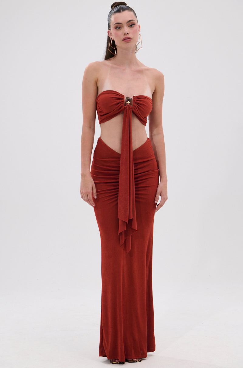 A woman models the ON MY MIND SLINKY MAXI SKIRT, wearing a strapless rust-colored gown with ruching, a cutout midriff, and a slinky maxi silhouette. Her hair is pulled back and she accessorizes with large geometric earrings against a white background. 4