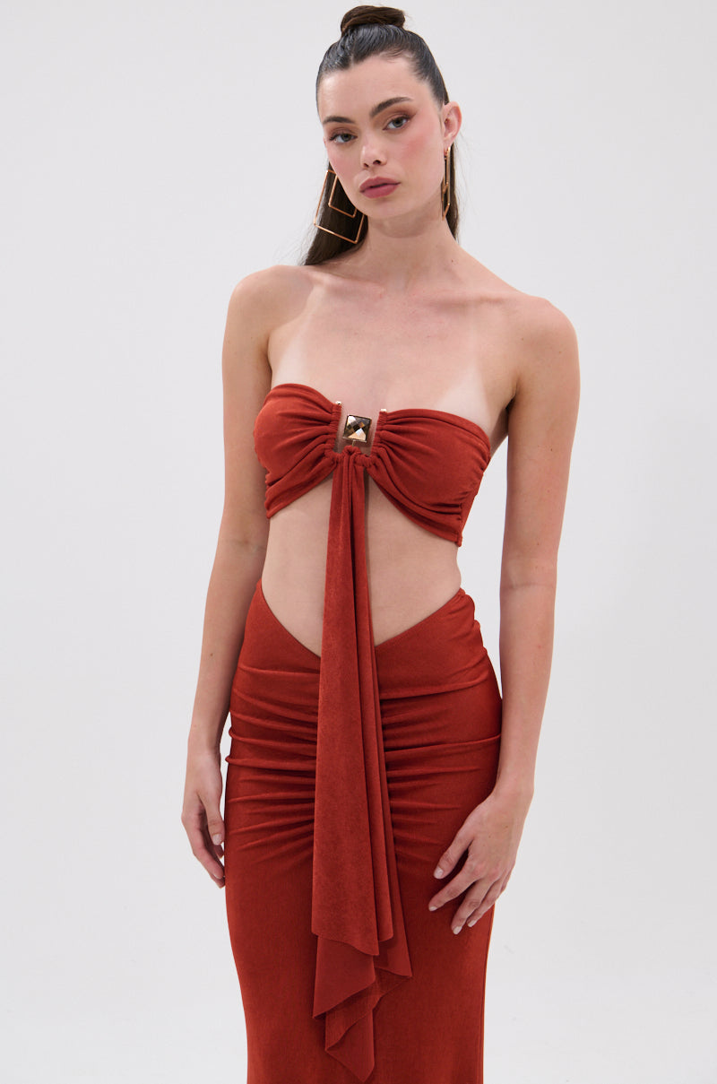 A woman models the ON MY MIND SLINKY BRALETTE, featuring a strapless neckline, rust color, front triangular cut-out with gold bust accent, and draped center fabric. She poses against a plain white background. 0