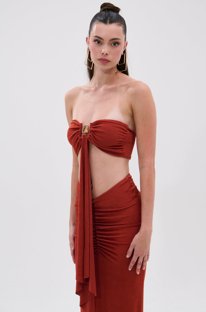 A woman with long dark hair in a high ponytail wears the ON MY MIND SLINKY BRALETTE, featuring a rust shade, strapless neckline, cut-out, and draped front tie. She stands against a white background, looking at the camera. 1