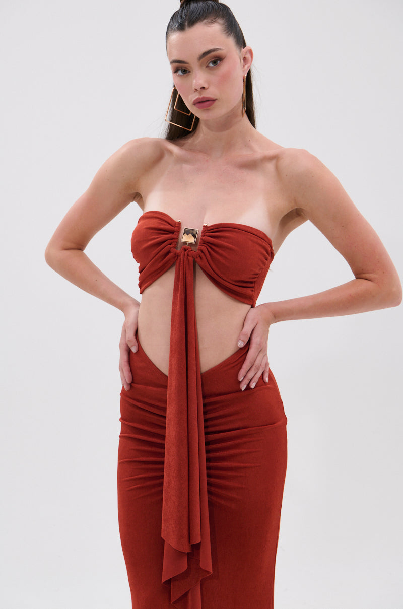 A woman poses with one hand on her hip against a plain white background, wearing the ON MY MIND SLINKY BRALETTE—a strapless, rust-red dress with a bralette-inspired neckline, front cutout, and draped fabric. 2