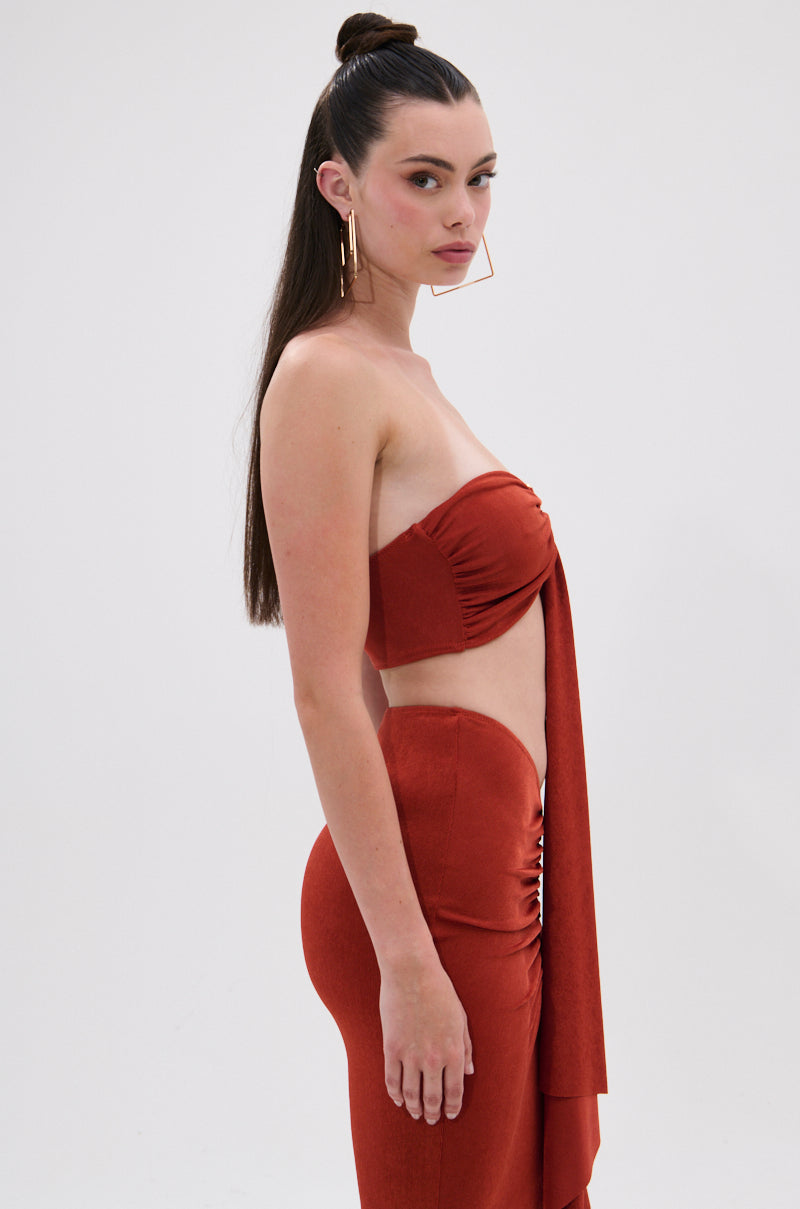 A woman with long dark hair in a high bun models the ON MY MIND SLINKY BRALETTE—rust-colored, strapless, with draped detail—paired with large geometric hoop earrings, shown in profile against a white background. 3