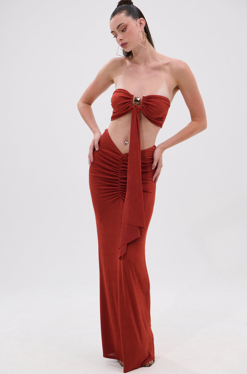 A woman poses in the ON MY MIND SLINKY BRALETTE, a rust-colored, strapless floor-length dress with front ruching and a chic central sash tie, accessorized with geometric earrings and slicked-back hair against a plain white background. 5