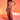 A woman stands against a red-orange gradient background wearing the DAYDREAMING SLINKY HIGH LOW SKIRT, styled as a light brown ruched two-piece with a crop top, sheer side panels, statement necklace, and strappy heels.