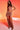 A woman stands against a red-orange gradient background wearing the DAYDREAMING SLINKY HIGH LOW SKIRT, styled as a light brown ruched two-piece with a crop top, sheer side panels, statement necklace, and strappy heels.