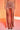 Wearing the DAYDREAMING SLINKY HIGH LOW SKIRT in ruched, high-waisted pink with two front slits, paired with black strappy high-heeled sandals, a person poses against a warm orange background.