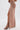 A person models the DAYDREAMING SLINKY HIGH LOW SKIRT, a high-waisted, ruched light brown maxi with a thigh-high slit, paired with strappy black heels against a plain light background.