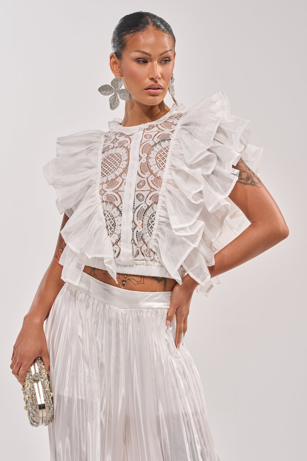 A woman models the BEST ONE YET LACE BLOUSE, featuring white lace and ruffled accents, styled with a pleated skirt and floral earrings. Hair pulled back, she holds a clutch—an ideal spring look against a simple background. 0