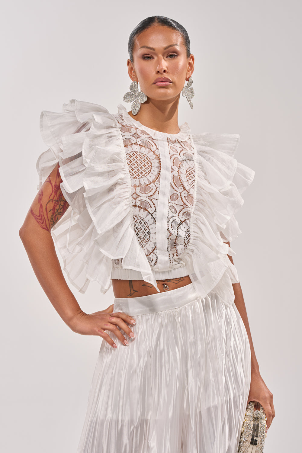 Wearing the BEST ONE YET LACE BLOUSE with large ruffled sleeves and a pleated white skirt, this confident individual pairs floral earrings, visible tattoos, and a small clutch for a fresh spring look. 1