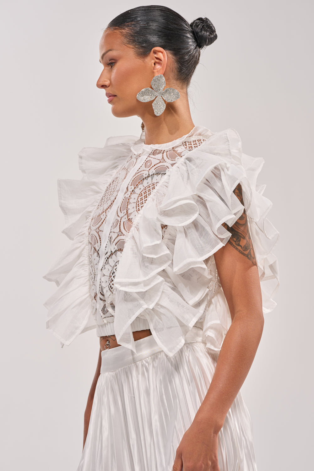 A woman with a sleek bun wears the BEST ONE YET LACE BLOUSE over a sleeveless lace top, paired with a flowy white skirt and bold flower earrings. She faces left against a neutral background, showcasing chic spring style. 2