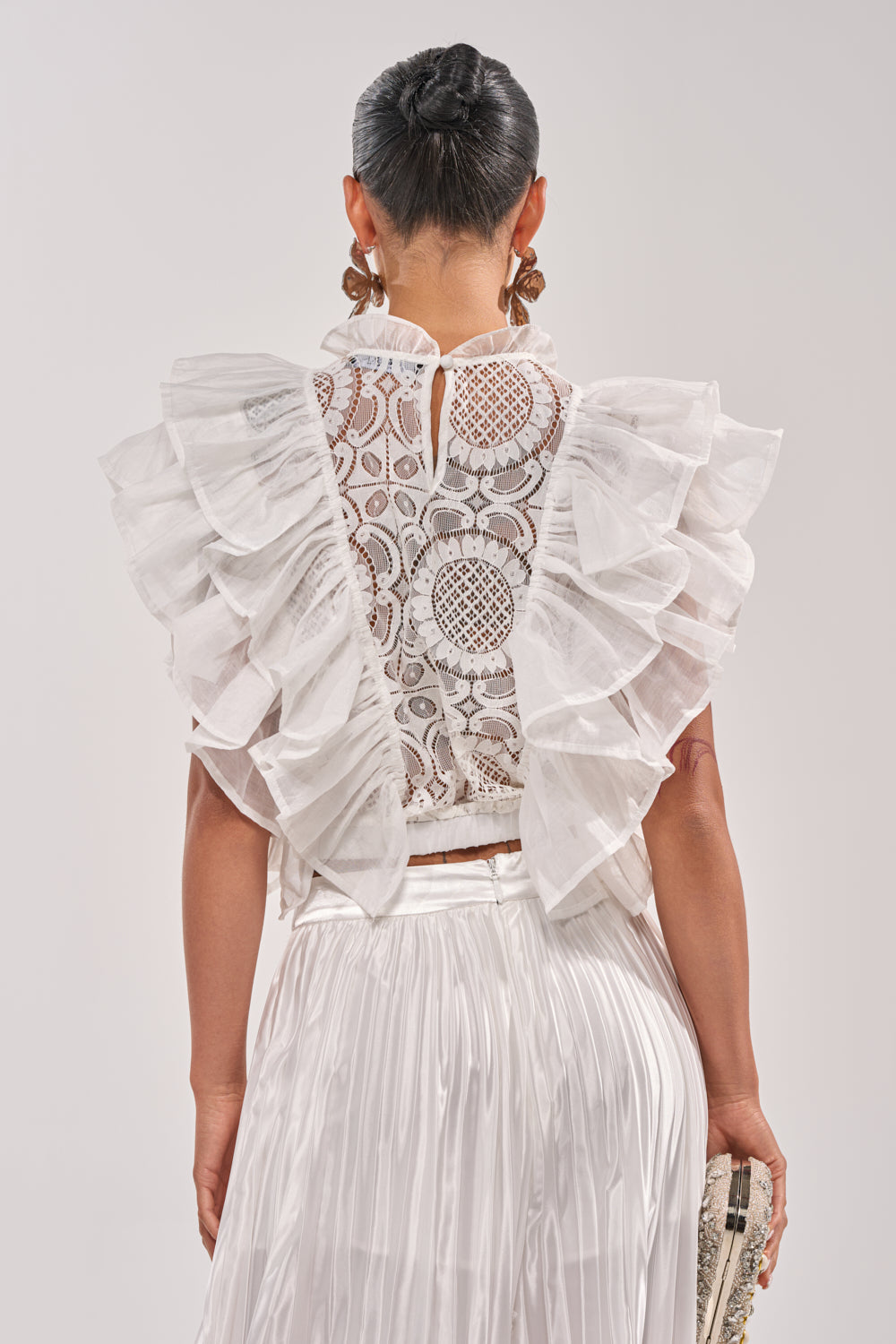 A woman models the BEST ONE YET LACE BLOUSE, featuring ruffled white lace with sheer circular patterns. Paired with a pleated white skirt and clutch, her chic spring look is completed with hair in a neat bun. 3