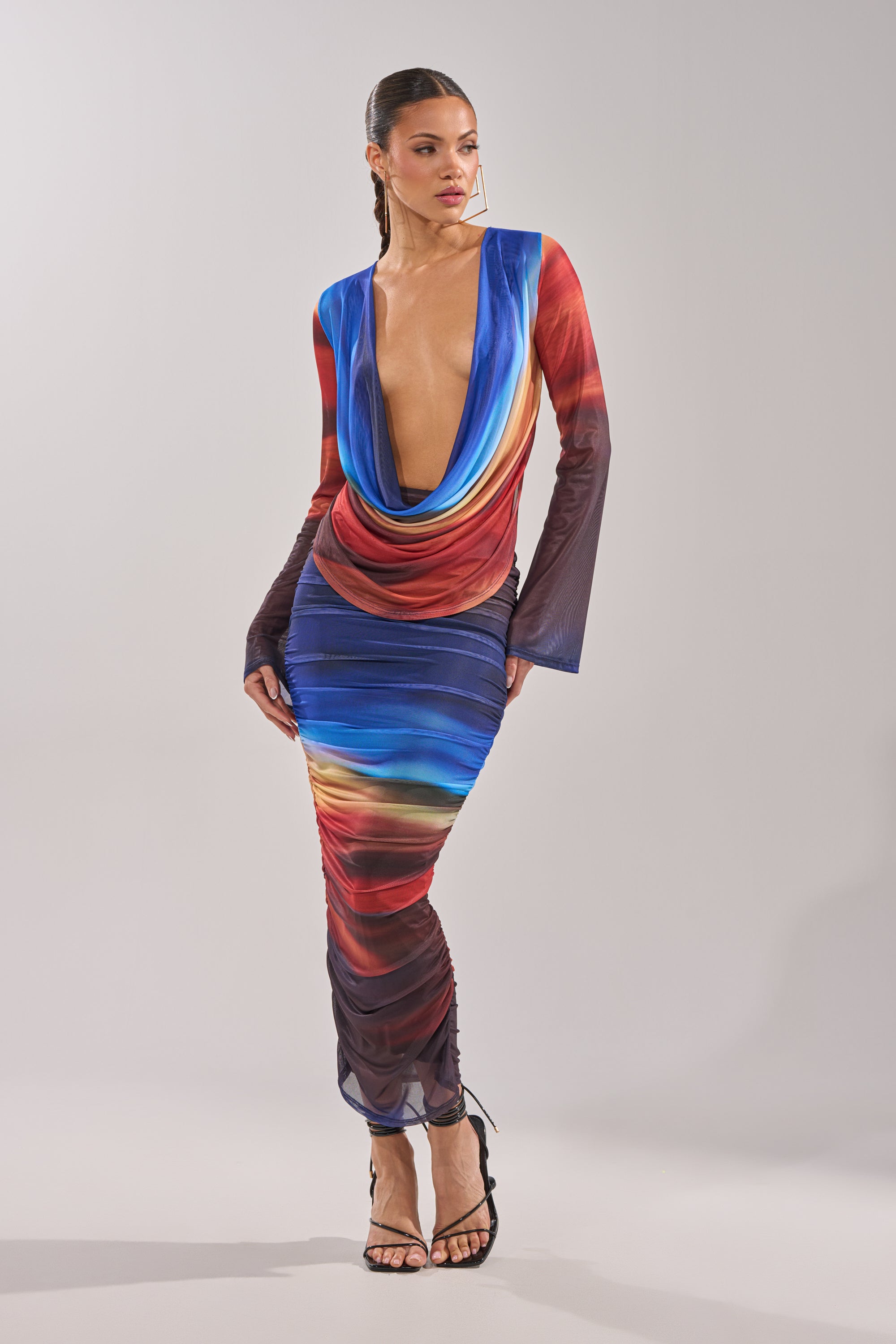 A woman wears the BOOM BOOM MESH MAXI SKIRT, a long ombre maxi skirt with ruched details and a vibrant blue, red, and yellow gradient. She pairs it with black strappy heels and has her hair styled back. 0