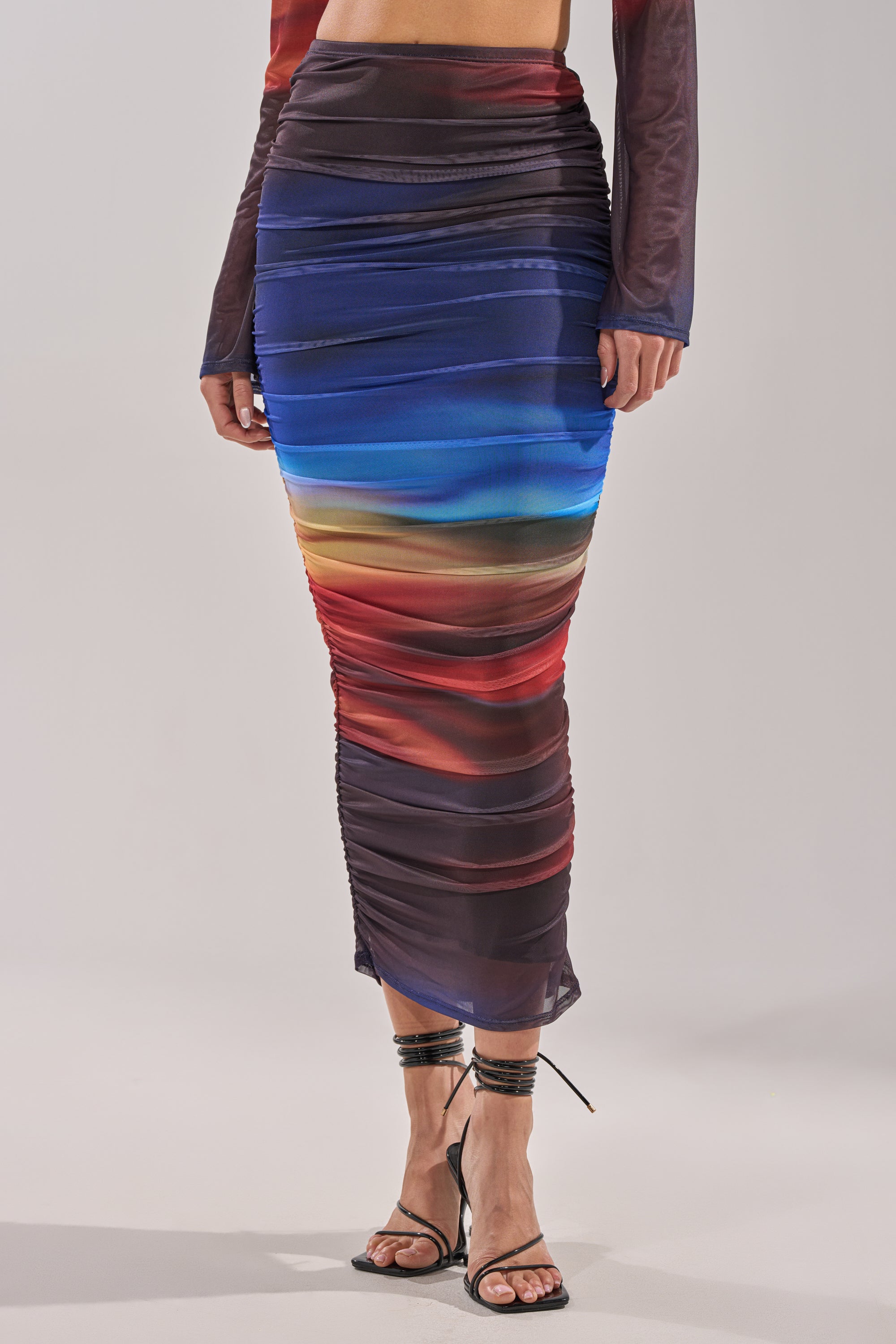 A person wearing the BOOM BOOM MESH MAXI SKIRT, a fitted high-waisted skirt in a blue, red, and brown sunset gradient, paired with strappy black heeled sandals and a matching long-sleeved top. 1