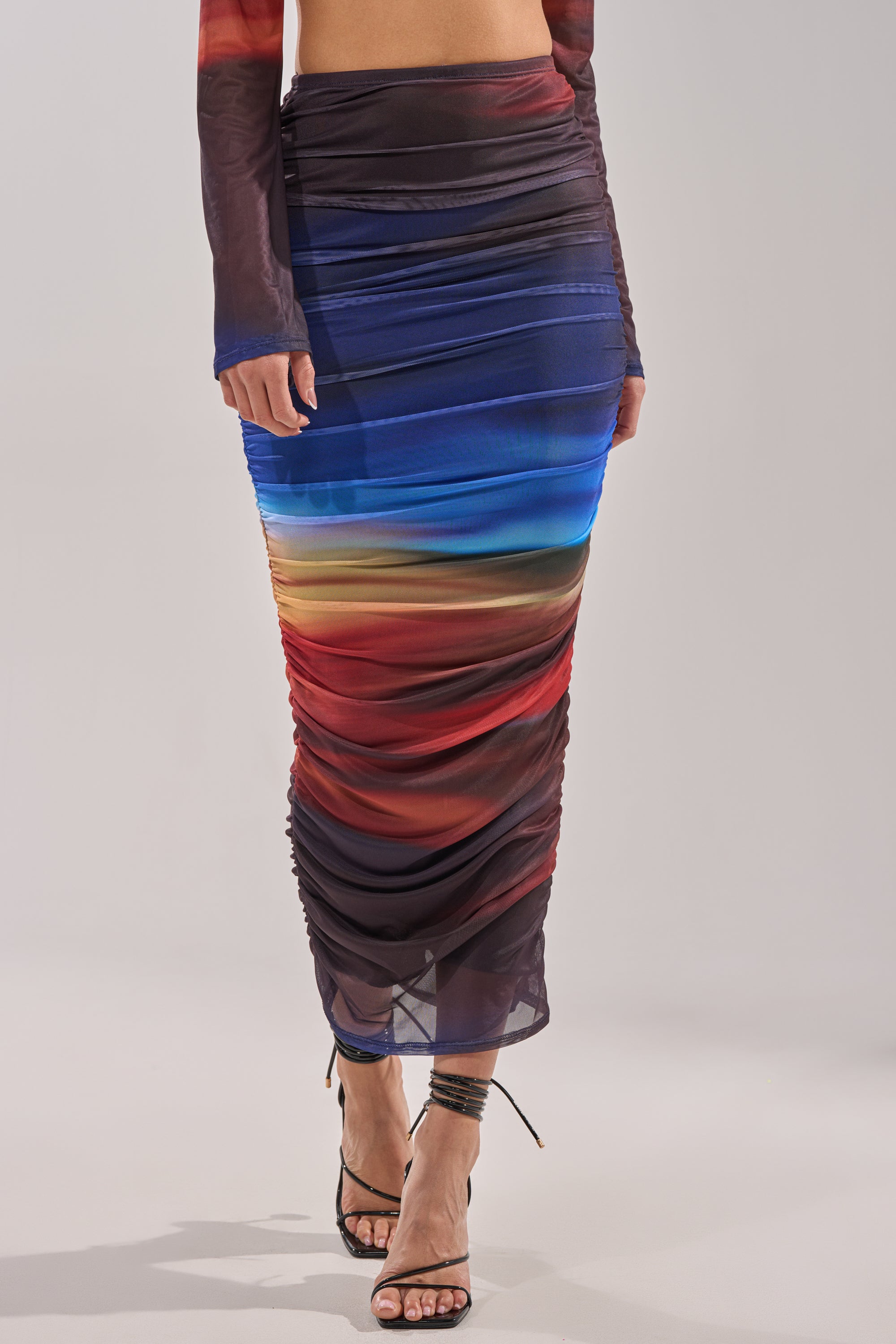 Someone wears the BOOM BOOM MESH MAXI SKIRT, featuring a ruched ombre design in red, blue, and orange sunset hues, paired with strappy high-heeled sandals. 2