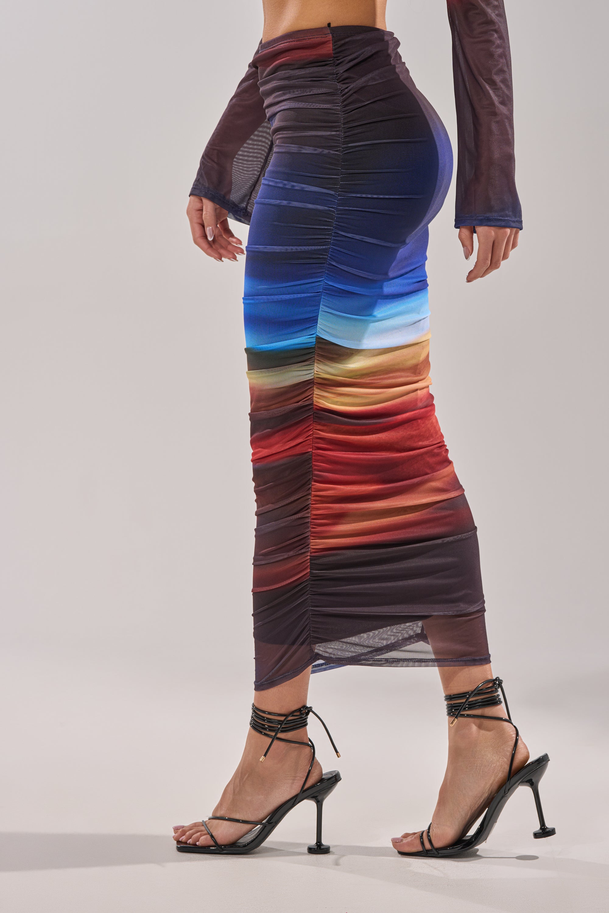 A woman wears black strappy heels and the BOOM BOOM MESH MAXI SKIRT, a fitted maxi with ruched ombre stripes in blue, white, brown, red, and purple. Her arms rest at her sides and her face is not shown. 3