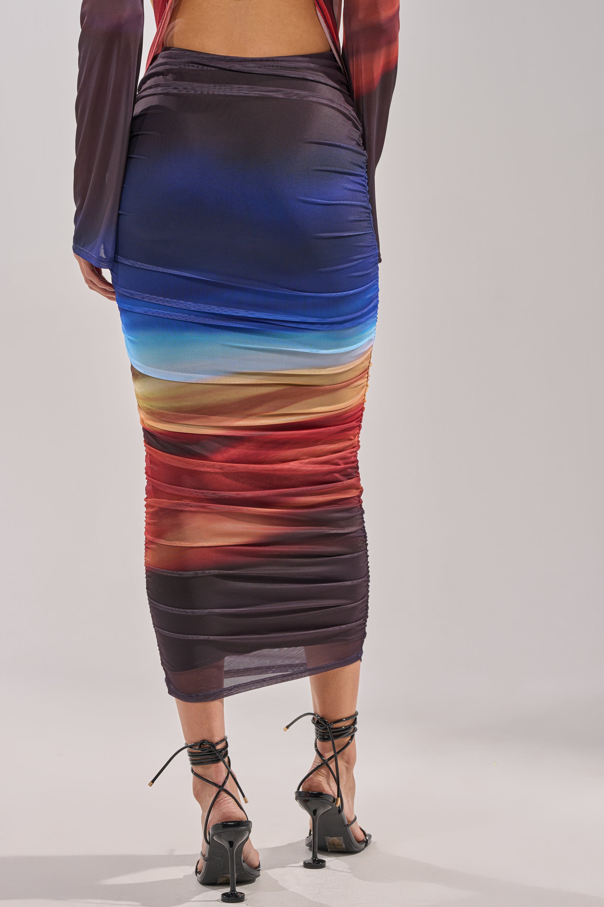 A person stands against a plain background wearing the BOOM BOOM MESH MAXI SKIRT, a fitted multicolored bodycon skirt with horizontal blue, red, brown, and beige stripes, paired with black strappy high-heeled sandals. 4