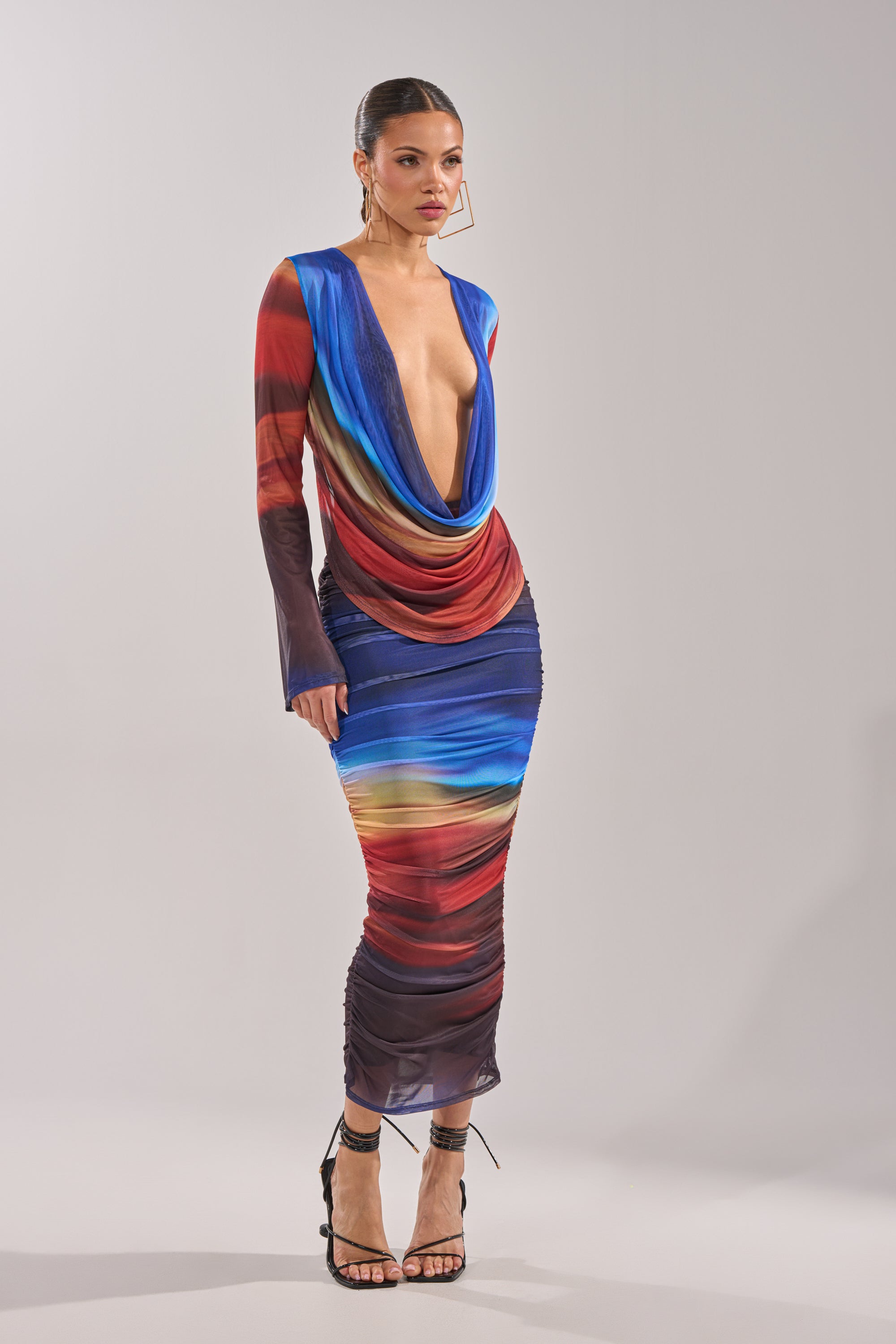 A woman poses against a plain background in the BOOM BOOM MESH MAXI SKIRT, styled with hoop earrings, black high-heeled sandals, and a sleek updo. 5