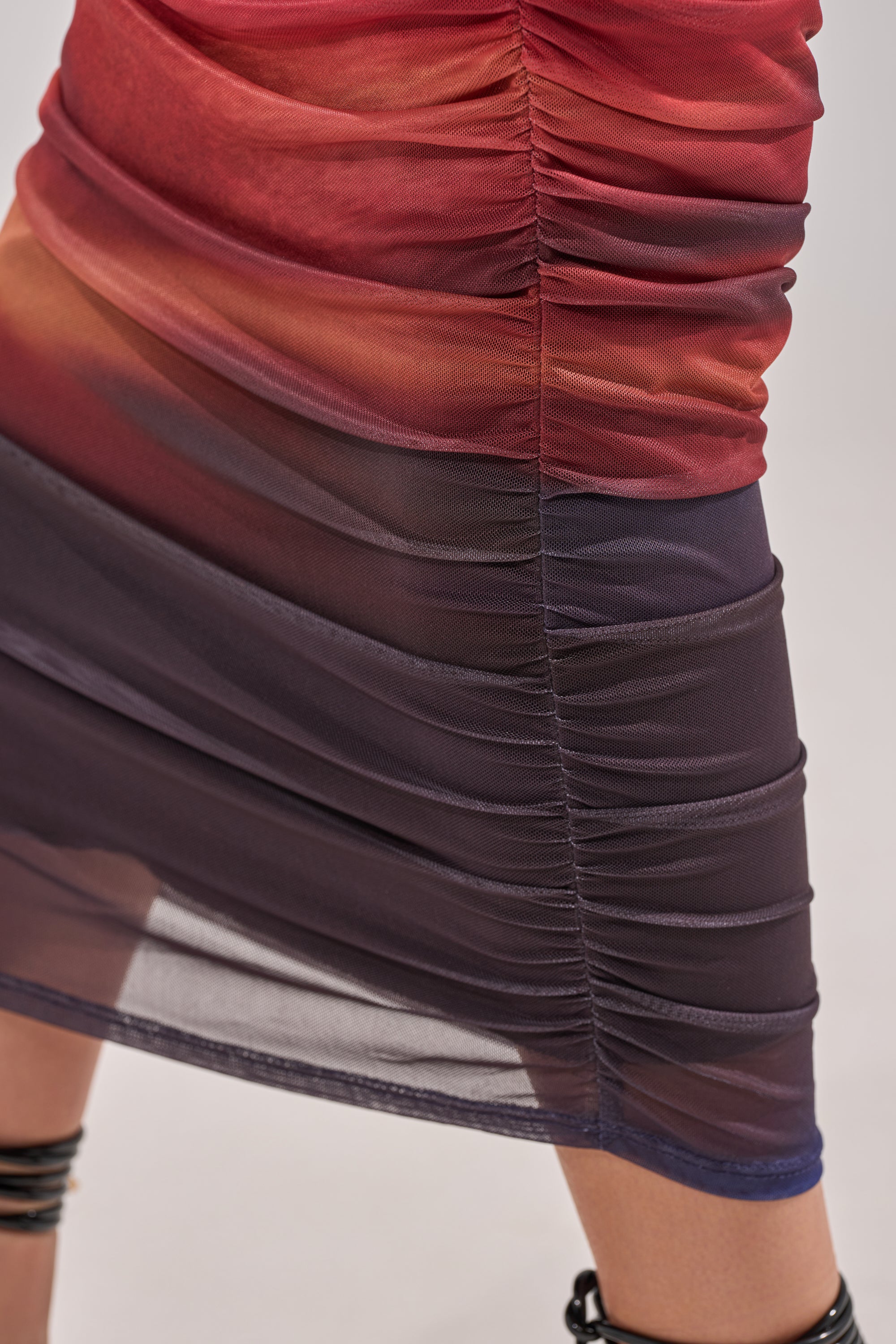 Close-up of the BOOM BOOM MESH MAXI SKIRT, featuring sheer mesh fabric in gradient red and purple ombre, ruched details, and a glimpse of a leg through gathered layers. 7