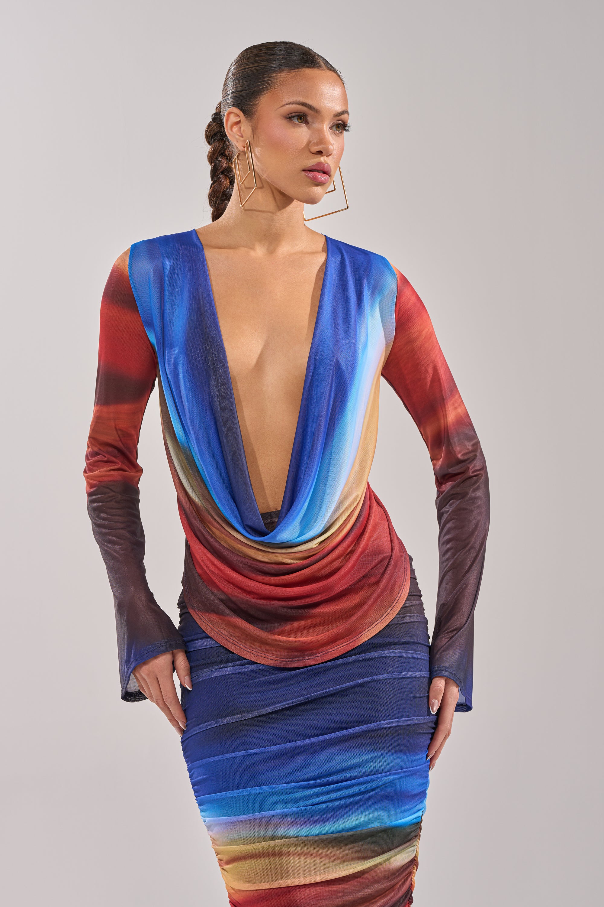 A woman with braided hair and large geometric earrings poses against a plain background, wearing the BOOM BOOM MESH BLOUSE—a long-sleeved, multicolored top with ombre mesh details in blue, red, and yellow and a deep cowl neckline. 0