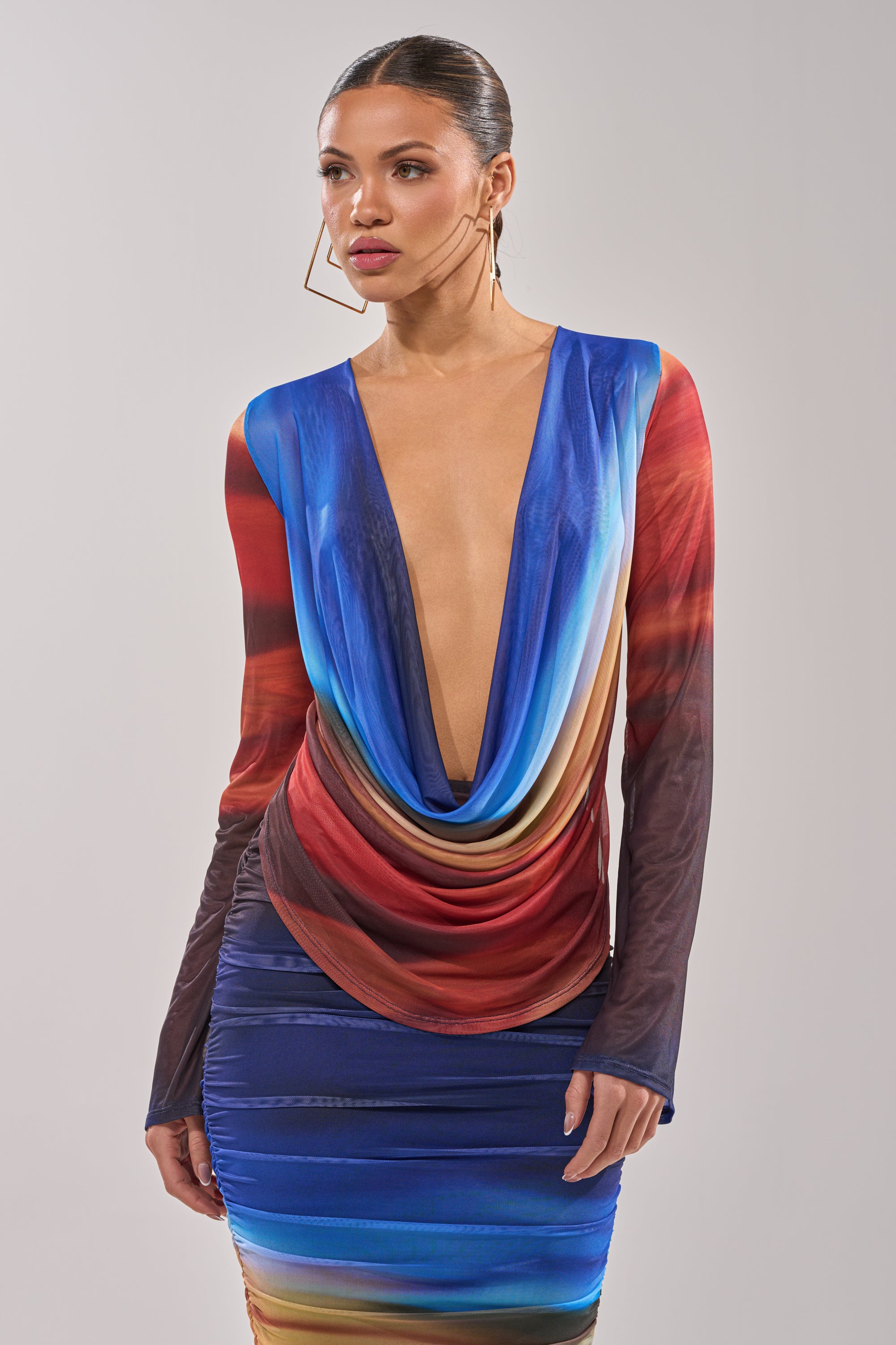 A woman wears the BOOM BOOM MESH BLOUSE, a long-sleeved, multicolored mesh top with a deep cowl neckline and blue, red, and brown gradient pattern. Her hair is sleekly pulled back, paired with bold geometric earrings. 1