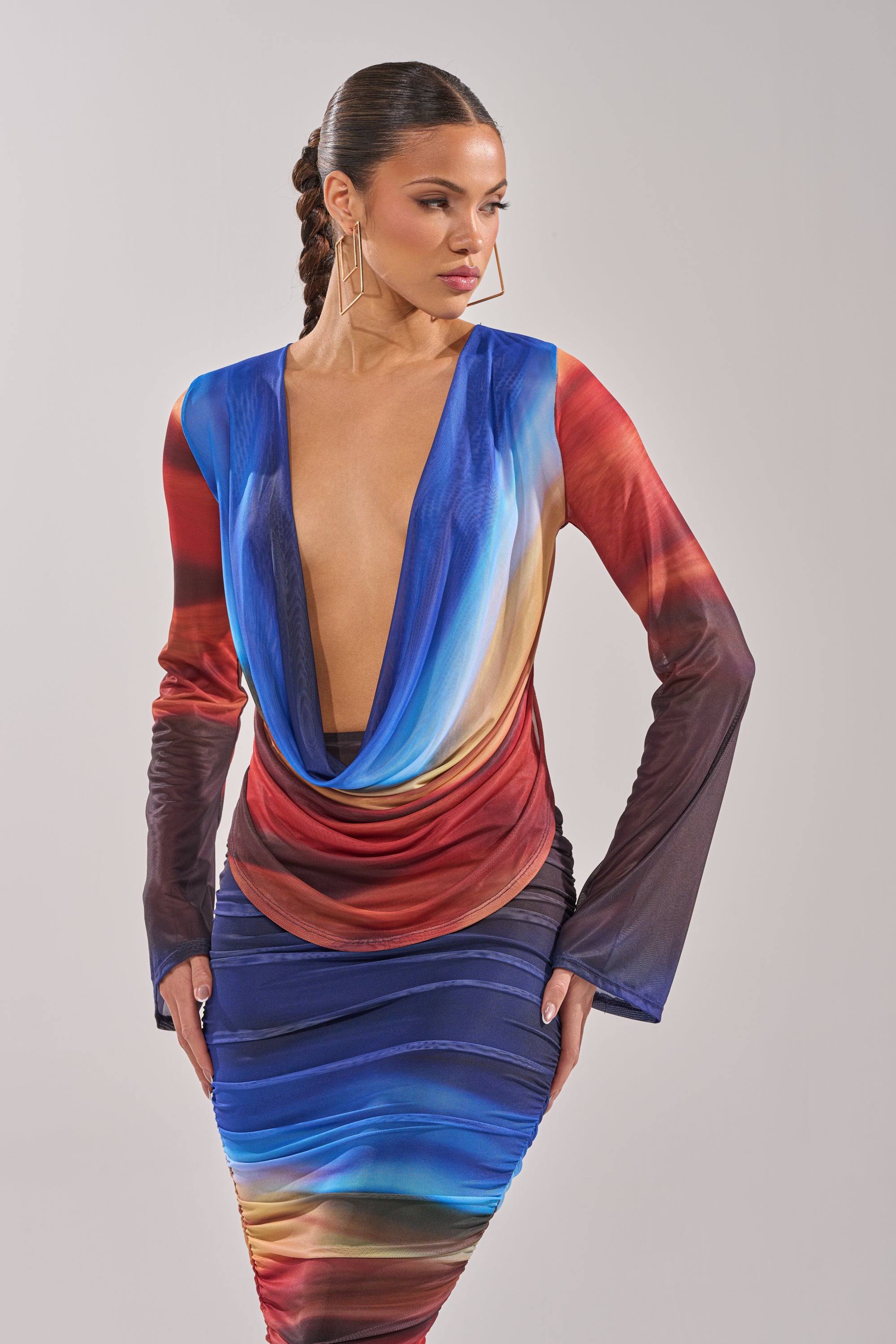 A woman in the BOOM BOOM MESH BLOUSE, a long-sleeved top with a deep cowl neckline and blue, red, and gold ombre mesh design, stands against a plain light background, gazing off to the side. 2