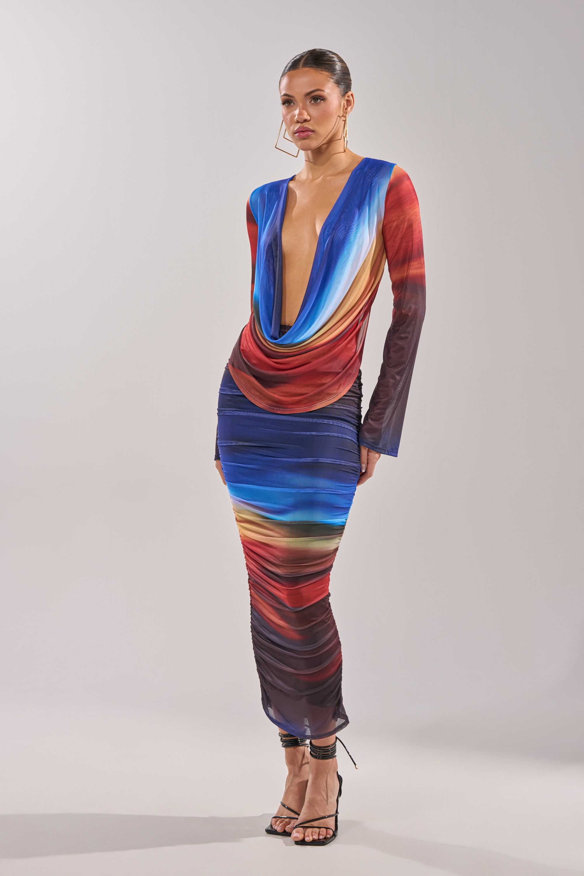 A woman wears the BOOM BOOM MESH BLOUSE, featuring long sleeves and vibrant ombre mesh stripes in blue, red, and yellow. She pairs it with strappy black heels, standing against a plain background. 7