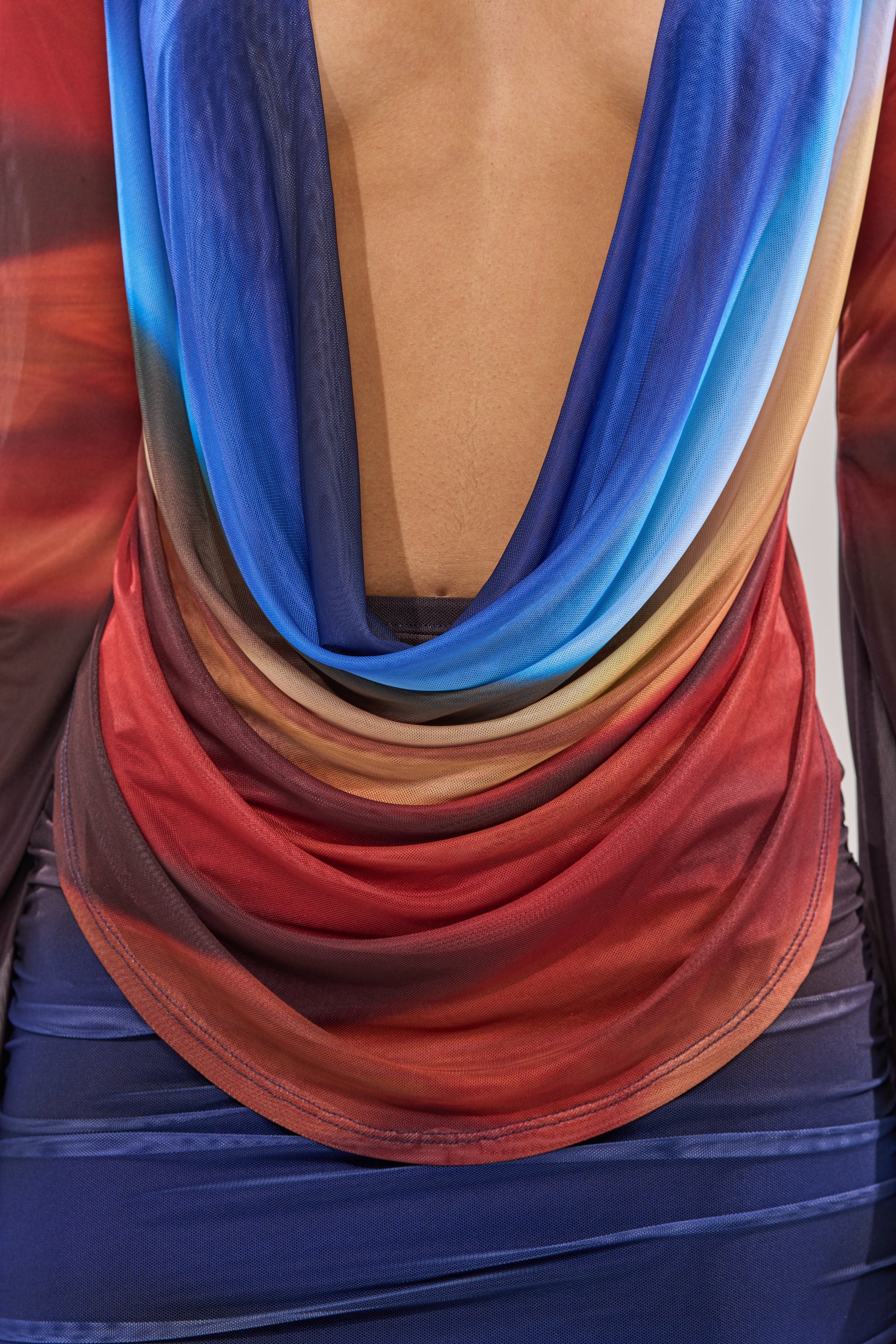A close-up of a person wearing the BOOM BOOM MESH BLOUSE, featuring sheer ombre mesh in blue, red, and tan with an open low back. The blouse is paired with a fitted blue skirt to complete the look. 8