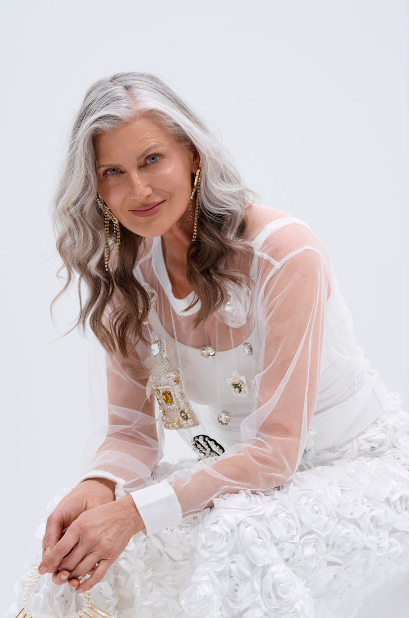 An older woman with long, wavy gray hair and gold earrings smiles gently against a plain background, wearing the GABRIELLA EMBELLISHED MESH TOP and a textured white skirt with delicate rosette details.