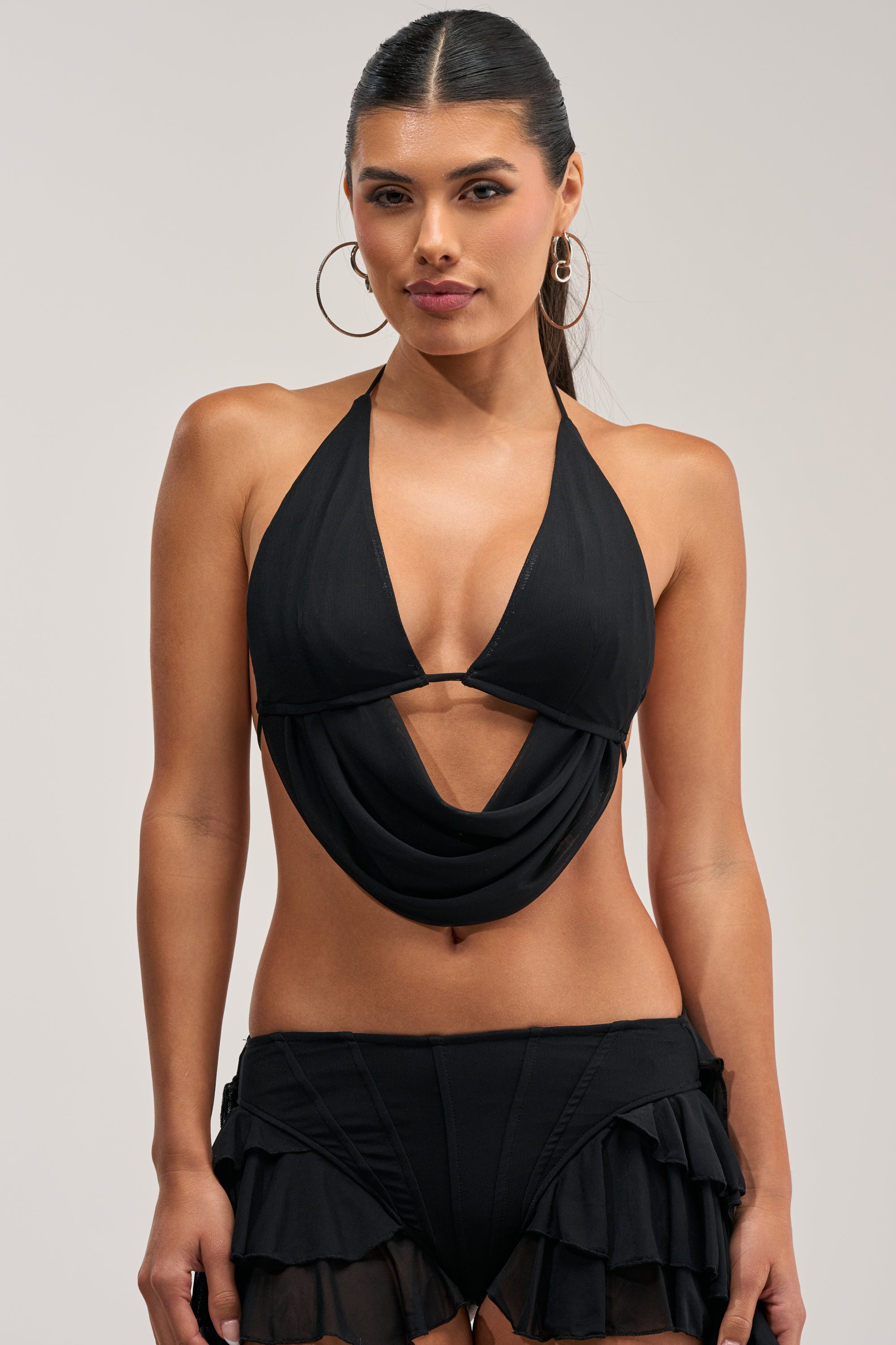 A woman wearing the FESTIVAL QUEEN BRALETTE TOP IN BLACK, featuring a stylish mesh halter design with a draped front, paired with matching black ruffled bikini bottoms—perfect for festivals or rave outfits—stands against a neutral background.