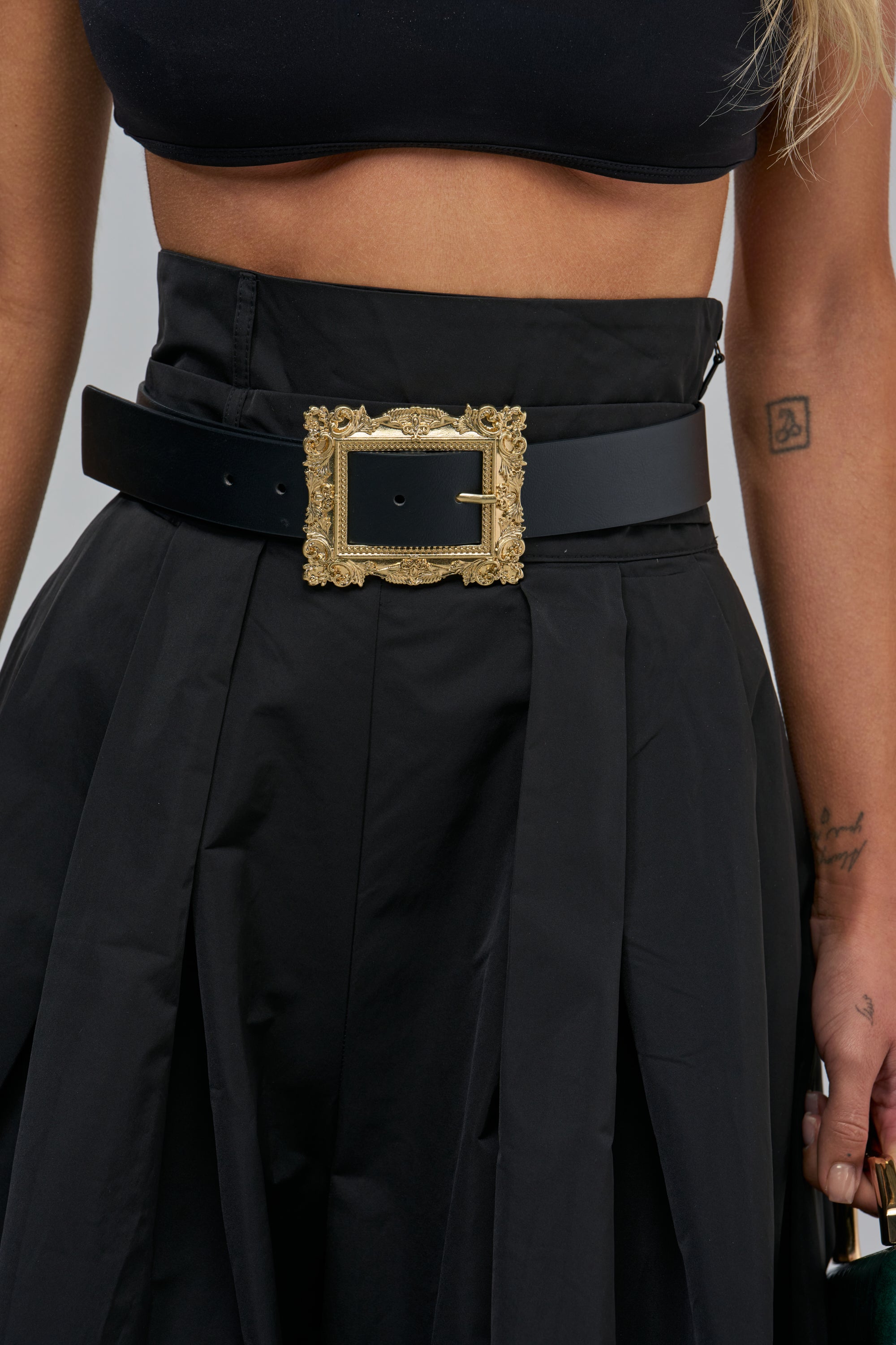 The STAY IN FRAME BELT, a black faux leather belt with a large, ornate gold buckle, worn over high-waisted black trousers.