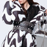 A woman showcases a fashionable ensemble wearing a black and white patterned puffer coat cinched with the LOVER GROMMET PU BELT, which features an adjustable metallic buckle. She accentuates the outfit with dark sunglasses and a metallic silver handbag for a sleek, contemporary style. Her straight brown hair adds the finishing touch to her look.